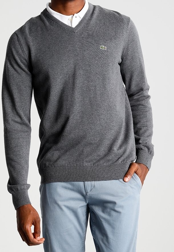 Homme portant un pull gris à col en V avec une chemise blanche à col et un pantalon bleu clair, main posée négligemment dans la poche.
