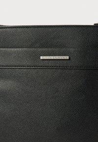 Melna teksturēta ādas soma ar sudraba metāla logotipa plāksni, uz kuras uzrakstīts "Armani Exchange", priekšpusē. Taisnstūra forma ar rāvējslēdzēja aizdari.