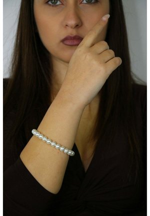 Pulsera de perlas con cuentas redondas y brillantes en blanco, ensartadas en un elástico. Se lleva en una muñeca sobre una prenda oscura. Punta del dedo apuntando hacia arriba.