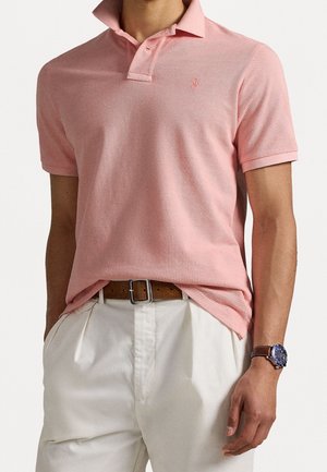 Polo rose pâle avec col, manches courtes et petit logo sur la poitrine, associé à un pantalon blanc et une ceinture marron.
