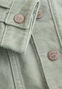 Tissu en denim vert clair avec des coutures détaillées et des boutons en métal étiquetés "Authentic 1982 Next Dude" sur une veste.