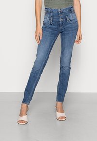 Jeans skinny, de cor azul escura, com um design de cintura alta, cinco bolsos e fechos de botão. Usados com sandálias de tiras de cor clara.