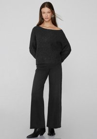 Junge Frau mit langen Haaren, die einen schulterfreien schwarzen Pullover, weite schwarze Hosen, schwarze Stiefel und silberne Ohrringe vor einem hellgrauen Hintergrund trägt.