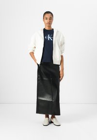 Felpa bianca con zip, t-shirt logo blu navy, gonna lunga in pelle nera e scarpe slip-on color crema. Texture lisce e palette di colori coerente.