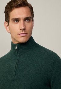 Dunkelgrüner Zip-Pullover mit geripptem Kragen, aus einem strukturierten Strickmaterial gefertigt, mit einem kleinen Logo auf der Brust.