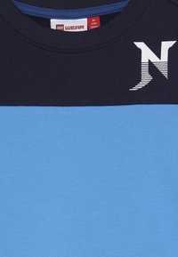 Chemise pour enfants avec un bloc de couleurs bleu marine et bleu clair, un "N" stylisé blanc sur la zone du col bleu marine, et étiquette de marque LEGO Wear pour les enfants de 4 ans.