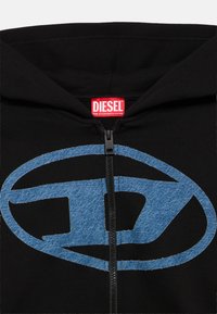 Μαύρο φούτερ με φερμουάρ, με μεγάλο μπλε τζιν λογότυπο στο μπροστινό μέρος και κόκκινη ετικέτα μάρκας στον λαιμό. Μαλακή υφή υφάσματος.