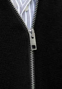 Sort strikket sweater med sølv lynlåsdetaljer, som har en stribet hvid skjorte under. Tæt på lynlåsområdet.