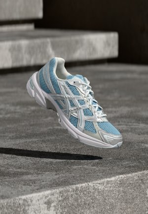 Zapatilla deportiva azul claro con parte superior de malla, acentos en blanco y suela acolchada. Presenta intrincados superpuestos y un sistema de cordones.