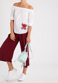 Haut blanc à épaules dénudées avec broderie florale rouge, associé à des culottes bordeaux et des baskets blanches. Portant un sac seau vert menthe.