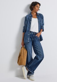 Blaue Jeanshemd, weißes Tanktop, blaue weit geschnittene Jeans, beigefarbene Wildledertasche, weiße Sneakers. Einfaches Design, lockere Passform, minimale Akzente.