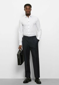 Weißes Hemd mit Knopfleiste und marineblauen Akzenten, kombiniert mit dunkler, maßgeschneiderter Hose. Eine schwarze Ledermappe und schwarze Schuhe vervollständigen den Look.