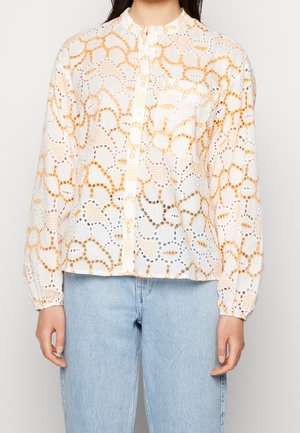 Femme portant un chemisier blanc à manches longues avec des motifs en dentelle orange et beige et un jean bleu clair sur un fond uni.