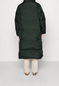 Lang, mørkegrøn pufferjakke med vandrette quiltede sektioner. Har en høj krave og sænkede skuldre. Båret over beige bukser og sandaler.