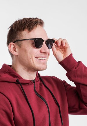 Round black sunglasses with dark lenses, silver metal accents, and a logo on the arm. Worn with a maroon zip-up hoodie.