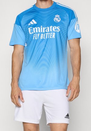 Muž v modrém dresu Realu Madrid s logem "Emirates Fly Better" a bílých šortkách Adidas stojí proti jednobarevnému pozadí.