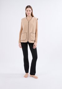 Gilet en polaire beige avec un col en V, deux poches plaquées et des bords contrastés à motif. Porté par-dessus un haut à manches longues et associé à un pantalon noir.