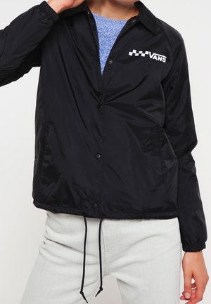Person iført en sort Vans skjortejakke over en blå sweater og hvide bukser, med snore hængende fra jakkeåbningen.
