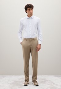 Camisa blanca de manga larga con botones y cuello, pantalones beige y zapatillas neutrales. La tela parece suave con un ajuste entallado.