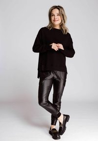 Schwarzer, übergroßer Pullover mit langen Ärmeln und seitlichem Schlitz, kombiniert mit engen schwarzen Kunstlederhosen mit Cargo-Taschen. Schwarze Plateau-Schuhe.