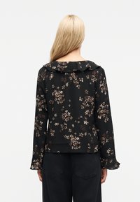 Γυναίκα με μακριά ξανθά μαλλιά φοράει μαύρη μπλούζα με λουλούδια, γιακά και μανσέτες με βολάν, συνδυασμένη με μαύρο παντελόνι, στέκεται μπροστά σε λευκό φόντο.