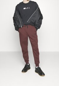 Černá dlouhá mikina s bílým logem Nike, černá bomber bunda, vínové tepláky s bočními kapsami a černé tenisky s béžovými podrážkami.