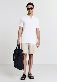 Polo bianco a maniche corte con colletto abbottonato, abbinato a pantaloni beige e sandali neri, mentre tiene una borsa a tracolla blu scuro.