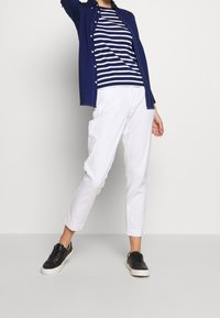 Person wearing white cropped pants, black sneakers, navy blue striped shirt, and open navy blue jacket standing against a plain light background.