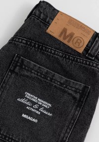 Zwarte denim jeans hebben een leren label met een geperst "M" en witte stiksels op het achterzakje met de tekst "MISAGA®" en andere details.