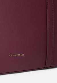 Bordó kézitáska struktúrált műbőrből, arany "ANNA FIELD" logóval és varrás részletekkel az oldalakon.
