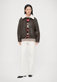 Brun bomberjacka i läder med vit pälskrage, har två framfickor och ribbade muddar, bärs över en randig tröja och vita byxor.