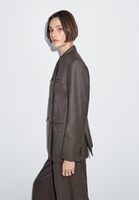 Hnědá saka na míru vyrobená z texturované tkaniny, s vysokým límcem, dvouřadým zapínáním a dvěma ozdobnými knoflíky.