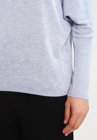 Maglione azzurro chiaro con un tessuto a maglia testurizzato, vestibilità morbida, polsini a coste e una sottile cucitura laterale. Abbinato a pantaloni neri.