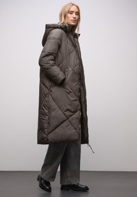 Manteau long matelassé gris foncé avec capuche, doté d'un motif matelassé, d'une fermeture éclair à l'avant et de poches latérales. Porté avec un pantalon gris à jambes larges.