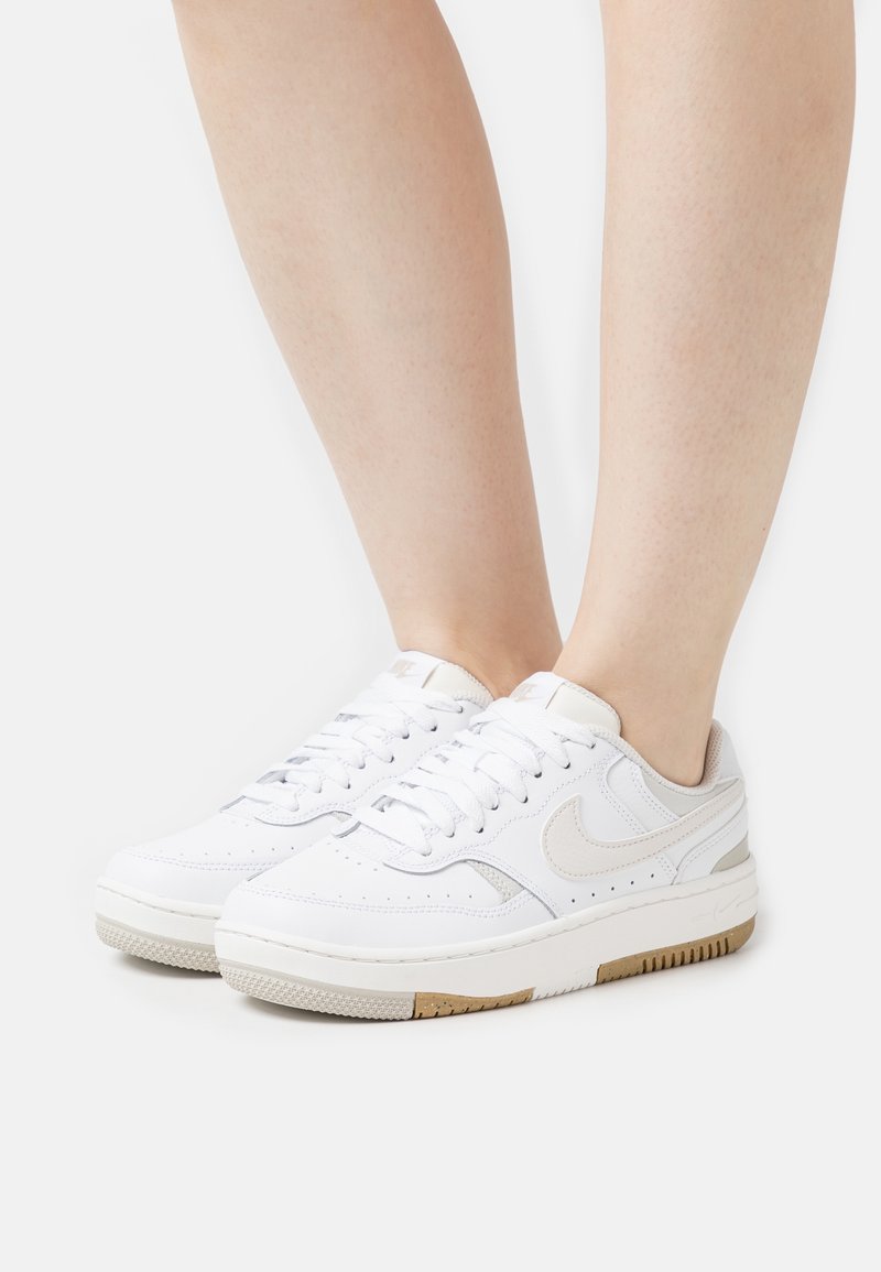 Jambes portant des baskets basses blanches Nike avec un swoosh gris clair et des accents de semelle couleur gomme sur un fond clair.