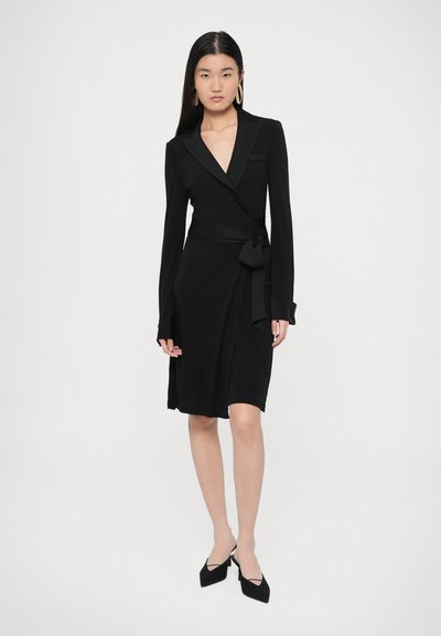 Μαύρο φόρεμα τύπου wrap με μακριά μανίκια, V-λαιμόκοψη και λεπτομέρεια δεμένη στη μέση. Απαλή ύφανση, λείο ύφασμα, μέχρι τους αστραγάλους, συνδυασμένο με μαύρα ψηλοτάκουνα.