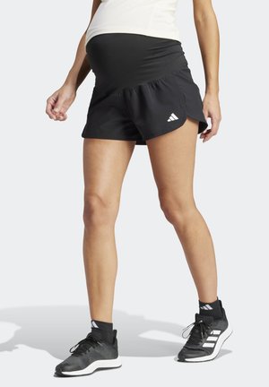Osoba w ciąży nosząca czarne sportowe krótkie spodenki ciążowe z logo Adidas, czarne skarpetki i czarne buty do biegania Adidas, stojąca na jasnym tle.