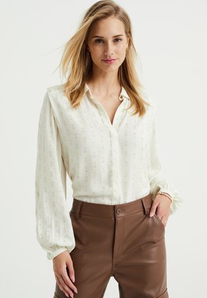 Witte blouse met knoopsluiting en een textuurpatroon, voorzien van puffmouwen, gecombineerd met bruine nep leren broeken. De blouse heeft een kraag en knopen.