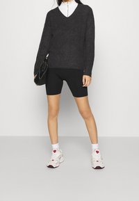 Maglione nero lavorato a maglia con scollo a V, abbinato a pantaloncini da bici neri e scarpe da ginnastica bianche. Accessori includono una borsa nera.