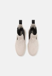 Béžové slip-on tenisky s čiernymi bočnými akcentmi, mäkká semišová textúra, slučka na päte a jednoduchý dizajn. Biela gumová podrážka.