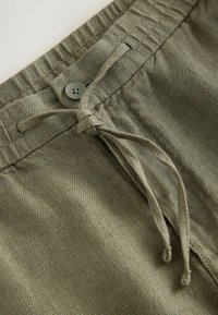Gros plan de la ceinture d’un pantalon vert olive avec élastique, cordon en tissu noué en nœud papillon, et un seul bouton vert au-dessus de la zone de la fermeture éclair.