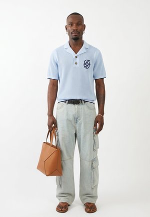 Uomo che indossa una polo a maglia azzurro chiaro, jeans cargo chiari, sandali marroni, che tiene una borsa a mano in pelle marrone con motivo geometrico, in piedi davanti a uno sfondo bianco.