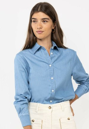 Mujer con cabello largo y castaño que lleva pendientes de aro dorados, una camisa azul abotonada y pantalones blancos, de pie con una mano en la cadera.