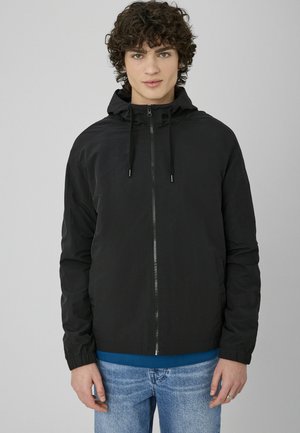 Jeune personne aux cheveux bouclés portant une veste noire à capuche zippée par-dessus une chemise bleue et un jean bleu clair, debout sur un fond uni.