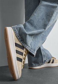 Μπεζ σουέτ αθλητικά παπούτσια με μαύρες ρίγες, κίτρινες κορδόνες και υφασμένη καφέ καουτσούκ σόλα. Τζιν παντελόνι με χαλαρή εφαρμογή ολοκληρώνει την εμφάνιση.