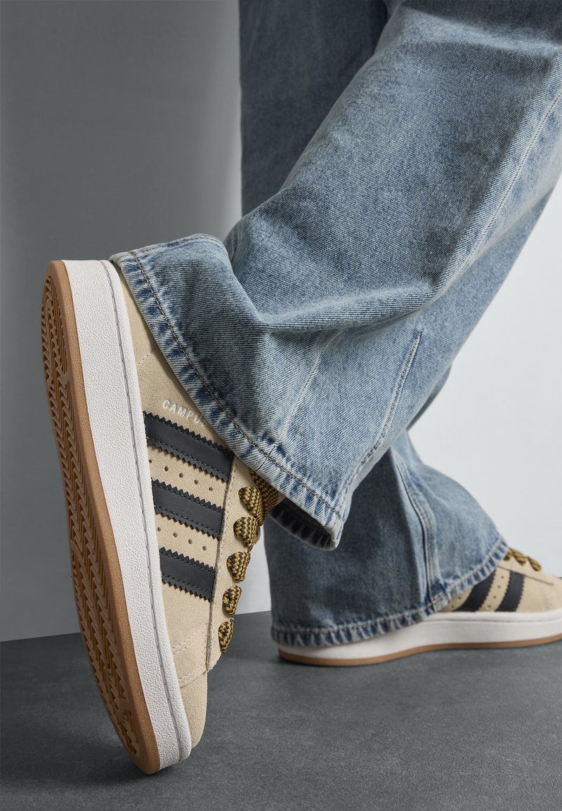 Μπεζ σουέτ αθλητικά παπούτσια με μαύρες ρίγες, κίτρινες κορδόνες και υφασμένη καφέ καουτσούκ σόλα. Τζιν παντελόνι με χαλαρή εφαρμογή ολοκληρώνει την εμφάνιση.