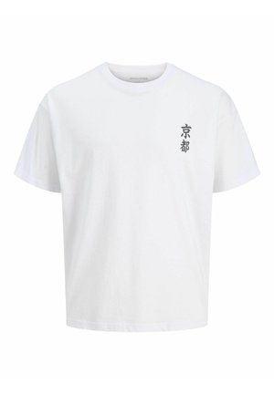 Garçon blond debout regardant à droite, portant un t-shirt blanc, un sweat à capuche bordeaux zippé, un pantalon beige ample et des chaussures claires.