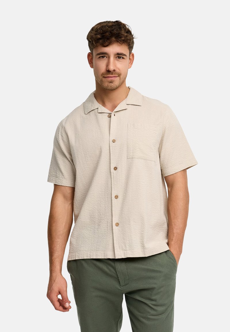 Man draagt een beige kortarmig getextureerd overhemd met knopen en een borstzak, gecombineerd met olijfgroene broek, tegen een effen achtergrond.