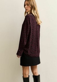 Camicia bordeaux con motivo a forma di cuore nero, maniche lunghe, design con colletto, indossata sopra una gonna nera e abbinata a stivali neri al ginocchio.