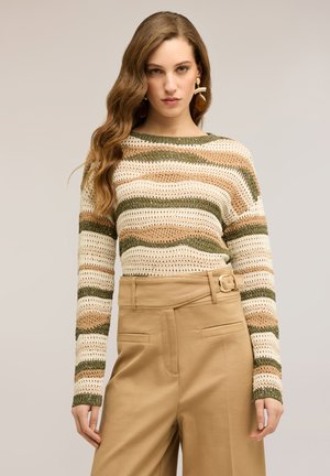 Junge Frau mit langen, welligen Haaren, die einen grün-beige-cremefarben gestreiften Strickpullover und hoch taillierte beige Hosen mit Gürtel-Detail trägt.